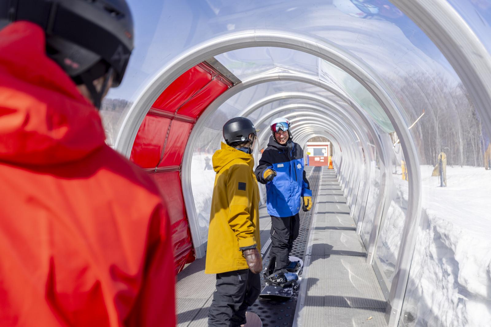 Snowboarders on a covered conveyor lift wearing helmets and colorful jackets.
