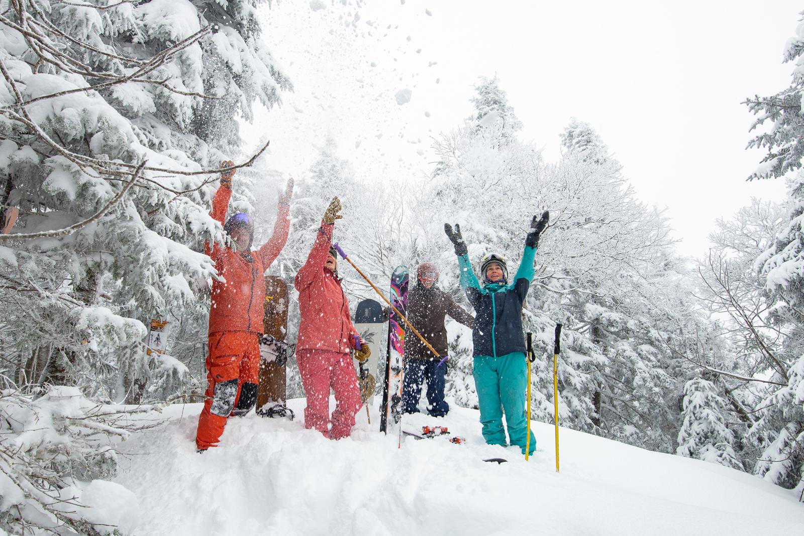Group of people in colorful ski gear joyfully throwing snow in a snowy forest.