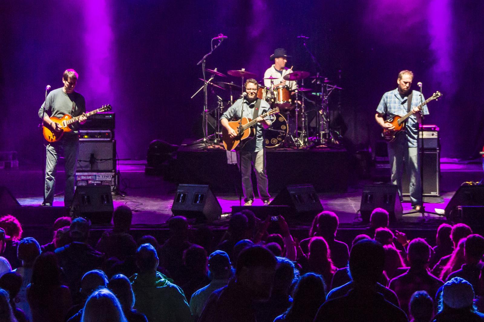 Rock band performing on stage with purple lighting, drum set, and guitars.
